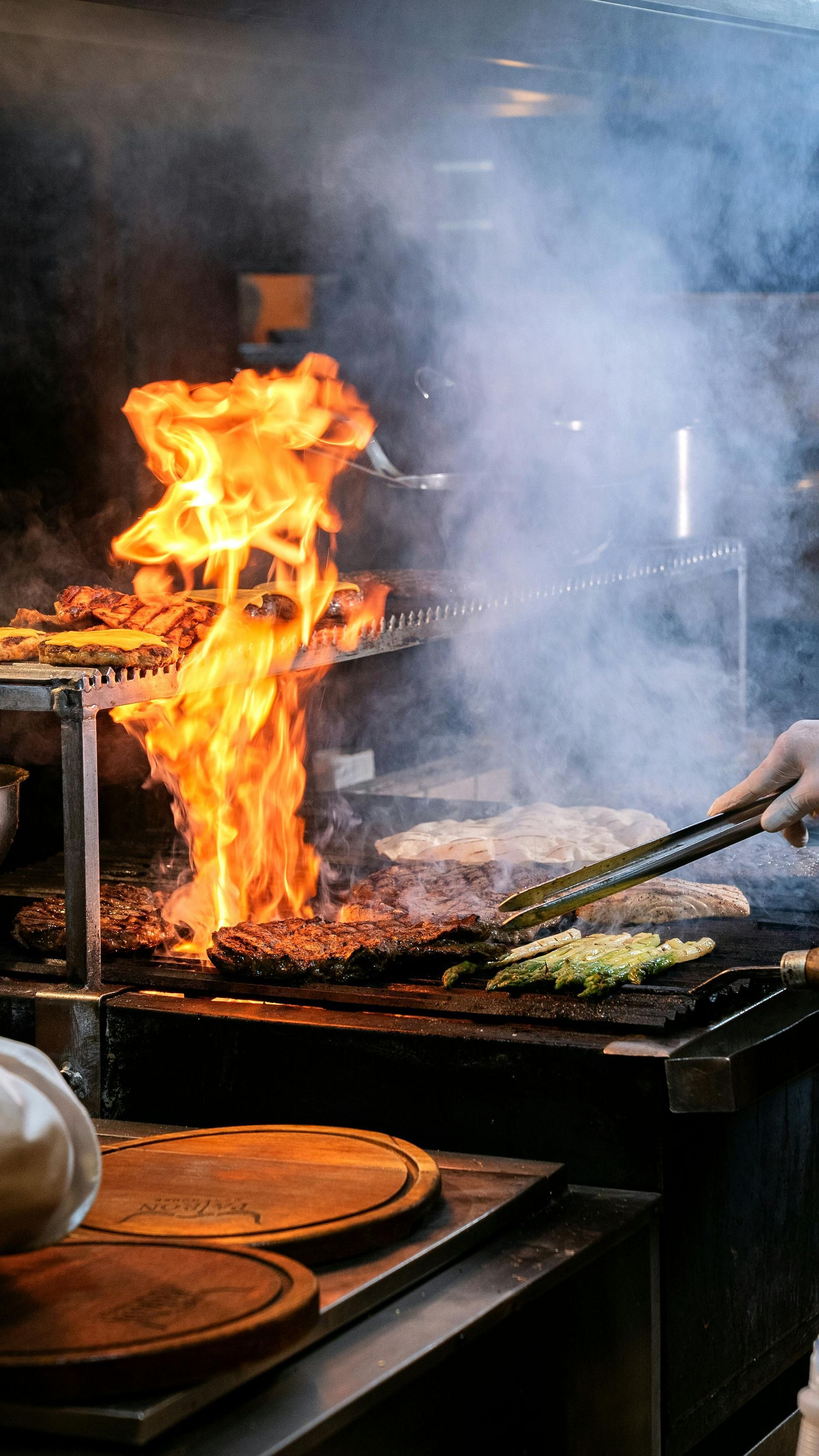 Chef flaming a pan in a smoky kitchen, with tongs grilling food on a stovetop.