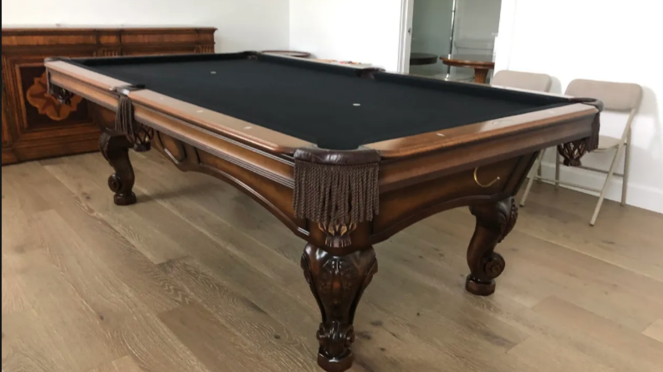 A wooden billiards table with a black felt surface, ornate carved legs, and fringed corner pockets in a room.