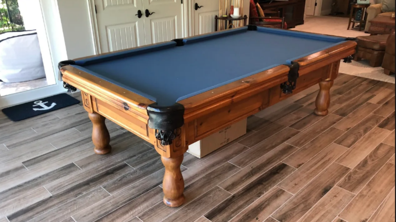 A wooden pool table with blue felt and black corner pockets stands on a light-colored wood-look tiled floor indoors.