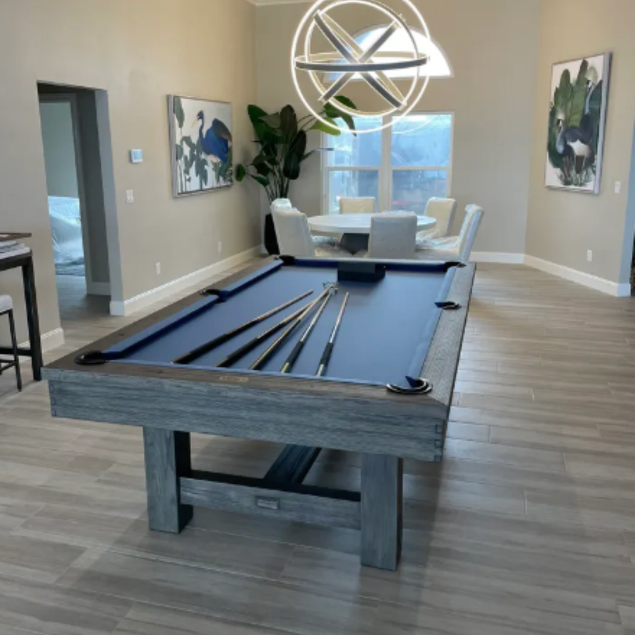 A pool table with blue felt sits in a modern, neutral-toned room featuring a round dining set and a spherical chandelier.