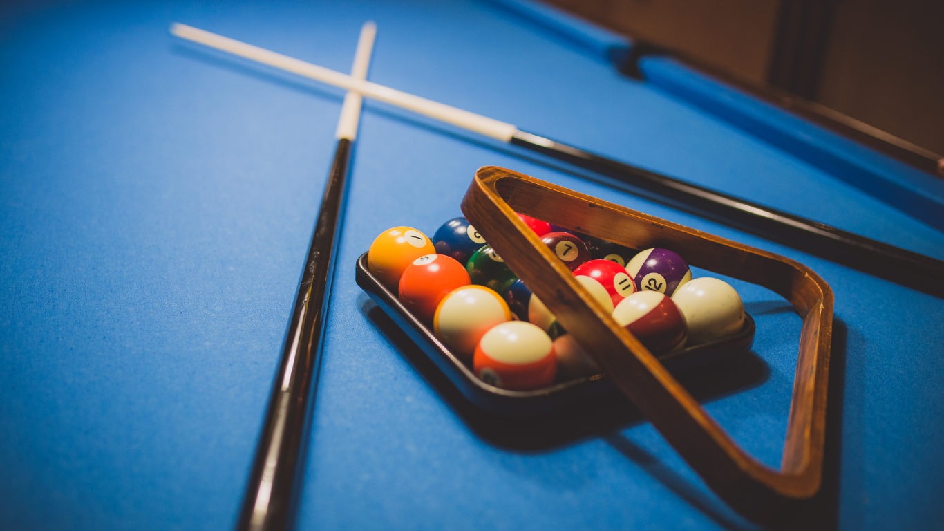 Two crossed pool cues rest on a blue pool table next to a triangle rack filled with colorful billiard balls.