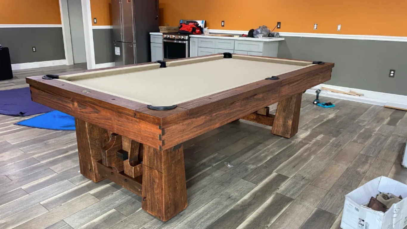 A rustic wooden pool table with a tan felt surface sits in a room with orange and gray walls and wood-look flooring.