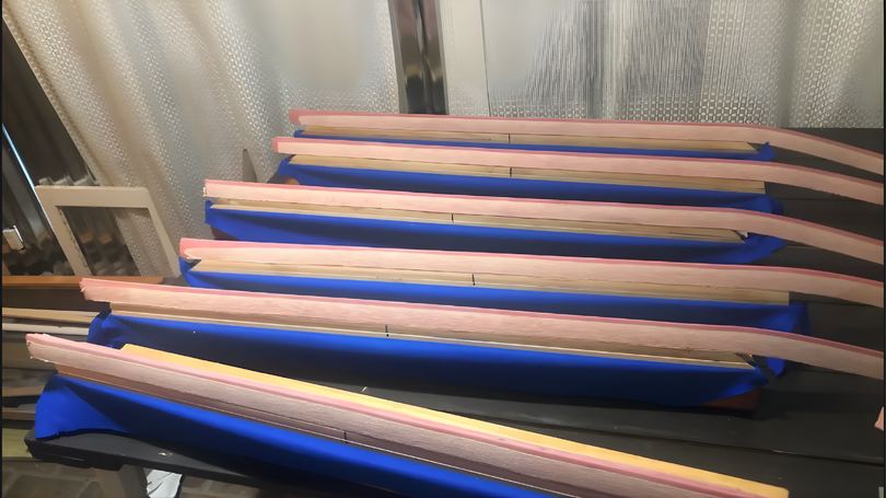 Four wooden guitar necks with frets resting on blue painters tape against a wall.