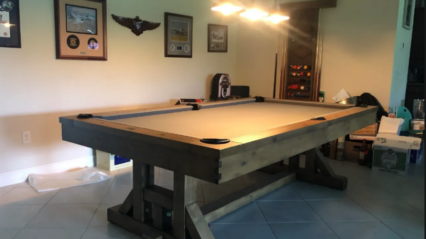 A rustic, gray-toned wooden pool table with a light-colored felt top sits in a room with framed decor on the wall.