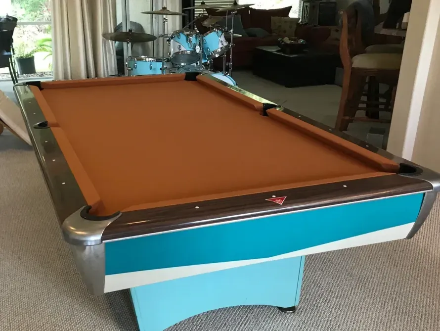 A teal and wood-toned pool table with a tan felt surface sits in a room with a drum kit in the background.