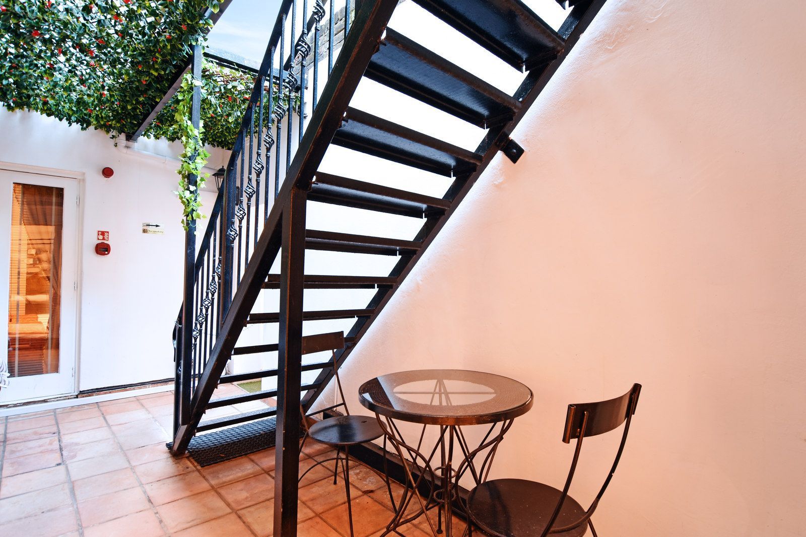 Black metal stairs with a small table and chairs in an outdoor courtyard with greenery.