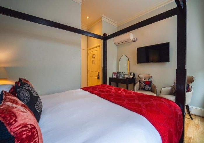 Bedroom with four-poster bed, red accents, TV, and vanity. Pale walls, dark frame, light wood floor.