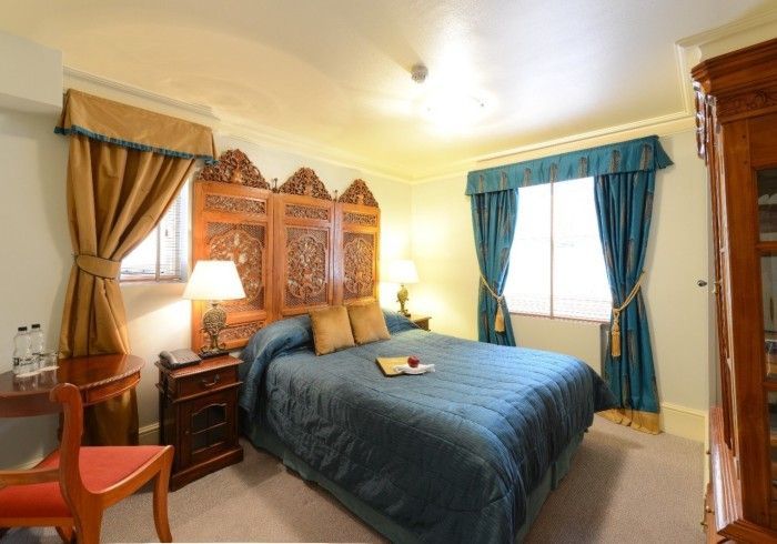 Hotel bedroom with ornate headboard, blue bedding, teal curtains, and wooden furniture.