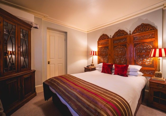 Cozy bedroom with ornate wooden headboard, double bed with red and brown accents, antique cabinet, and side tables.