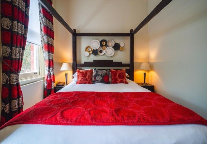 Bedroom with four-poster bed; red bedding and patterned curtains; white walls; lamps on nightstands