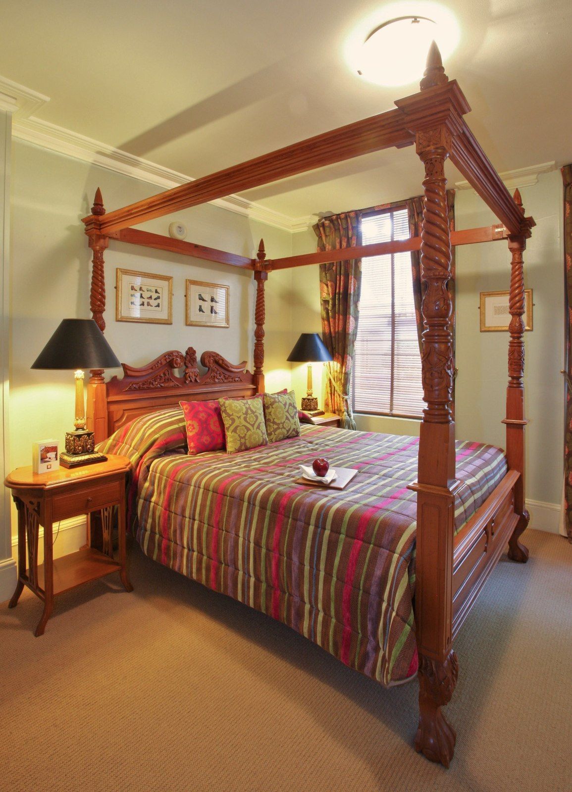 A bedroom with a large wooden canopy bed, side table with a lamp, and window with curtains.
