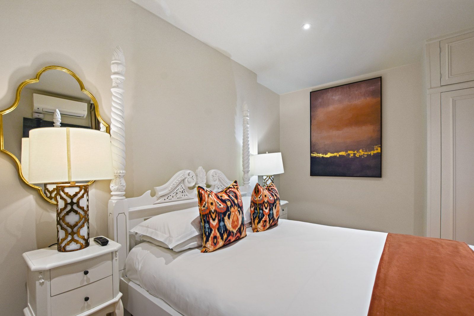 Bedroom with white bed, patterned pillows, gold mirror, orange accents, and artwork.