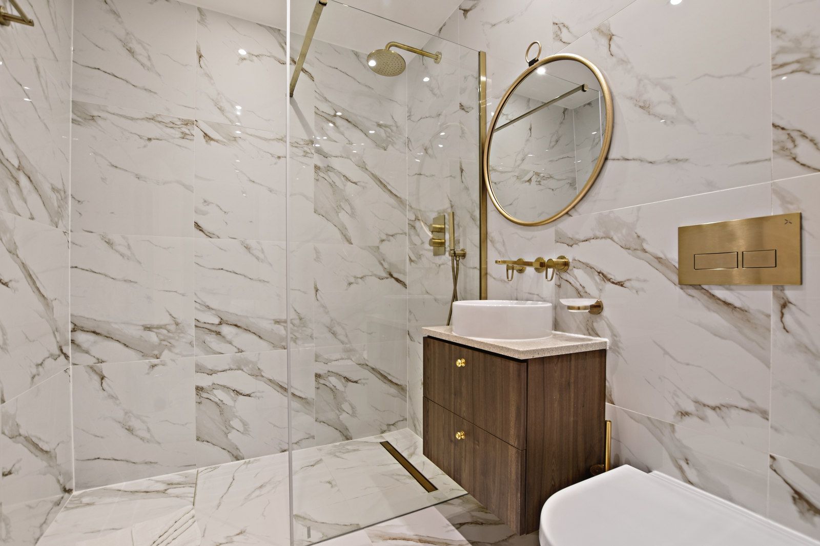 Luxurious bathroom with marble walls, gold fixtures, wood vanity, and a round mirror.
