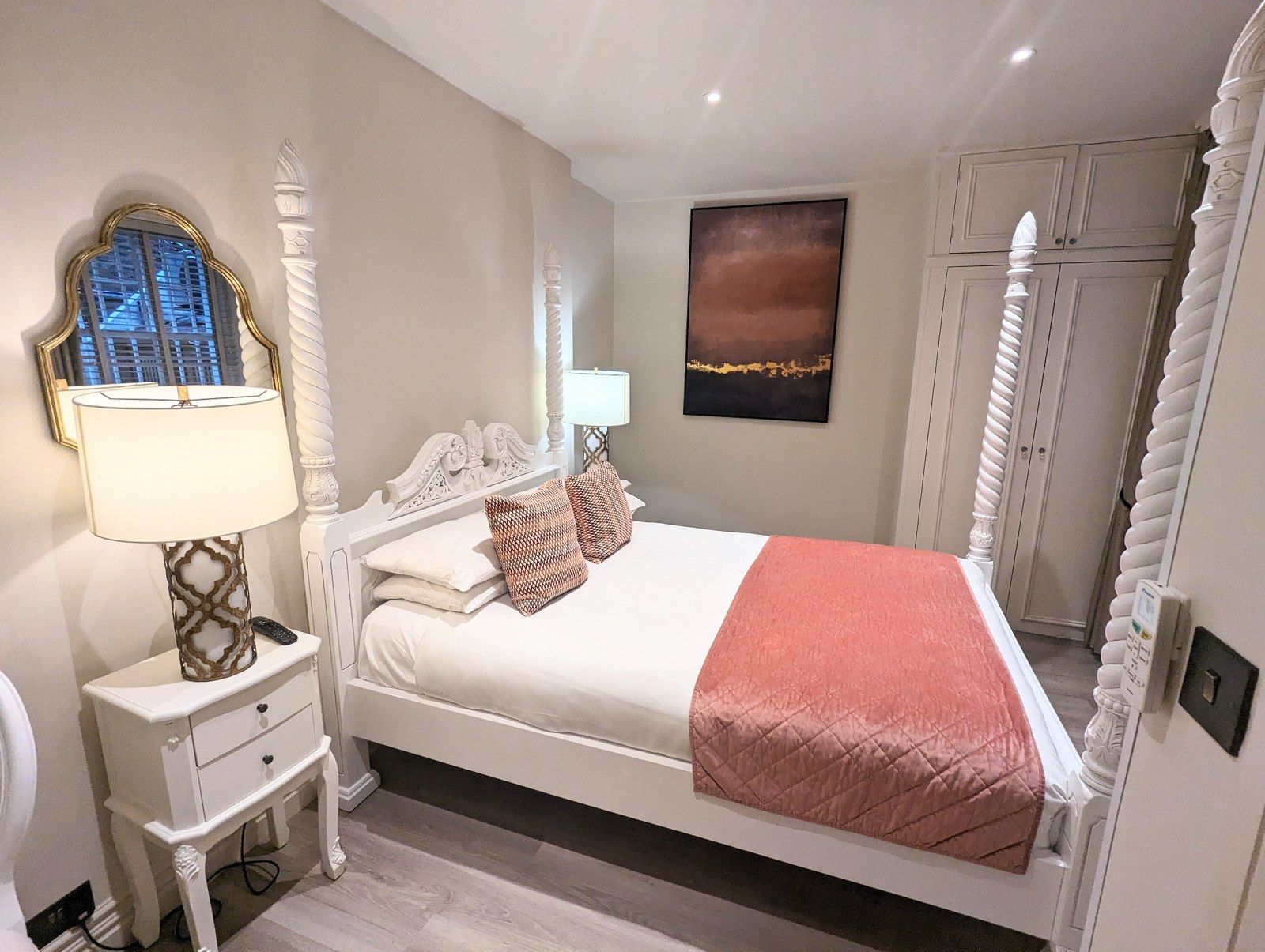Bedroom with white four-poster bed, nightstand, and wardrobe. Soft colors: white, peach, and gold.
