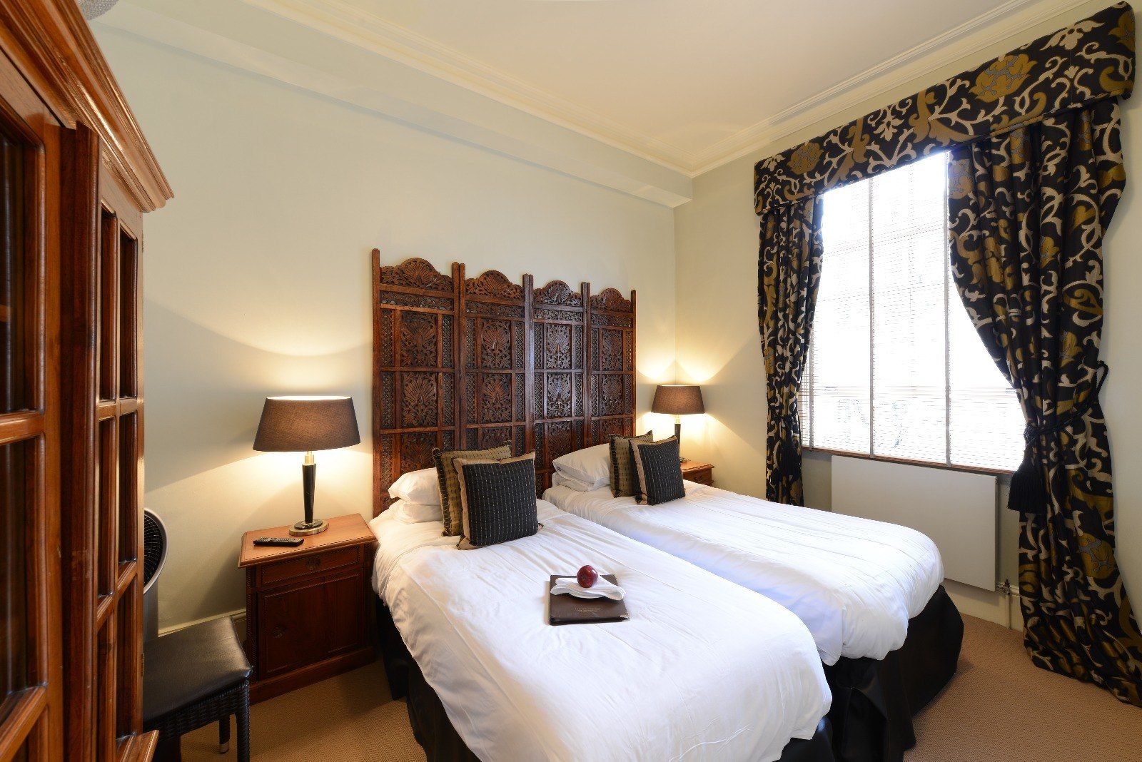 Two twin beds in a room with a decorative headboard, dark lamps, and patterned curtains.