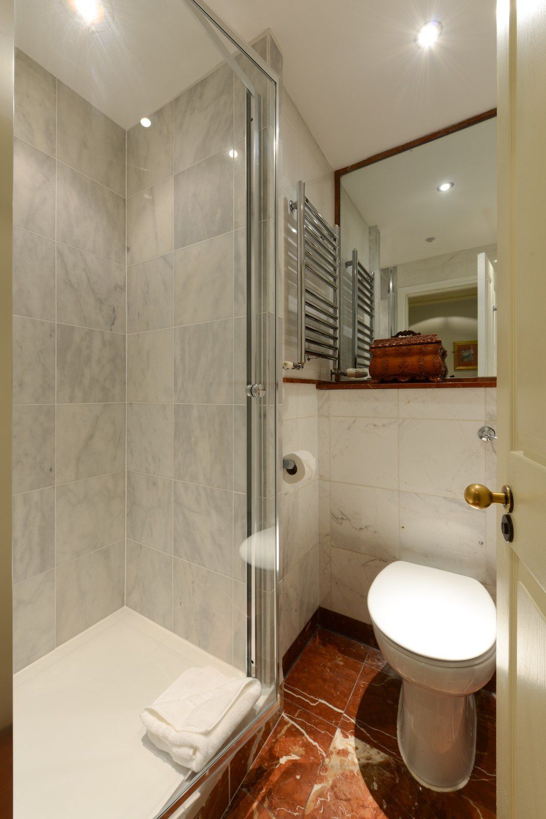 Small bathroom with marble shower, toilet, towel, and heated towel rack. Red floor.