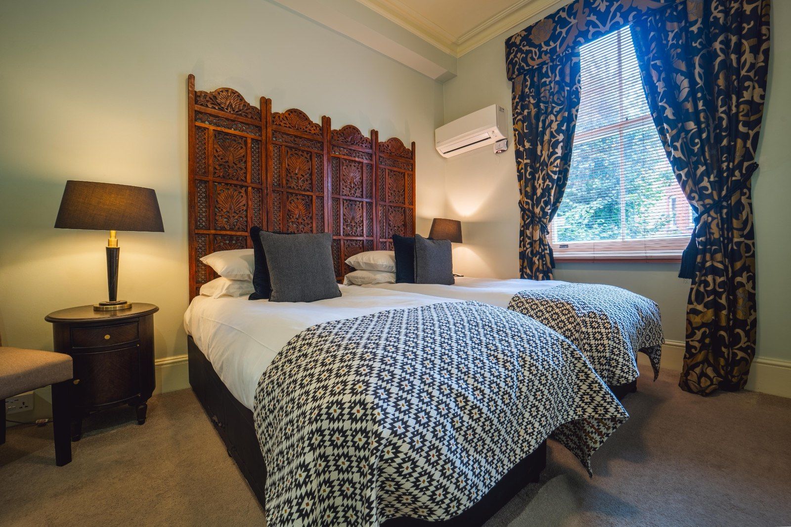 Twin beds with ornate headboard, dark throw blankets, table lamps, and floral curtains.