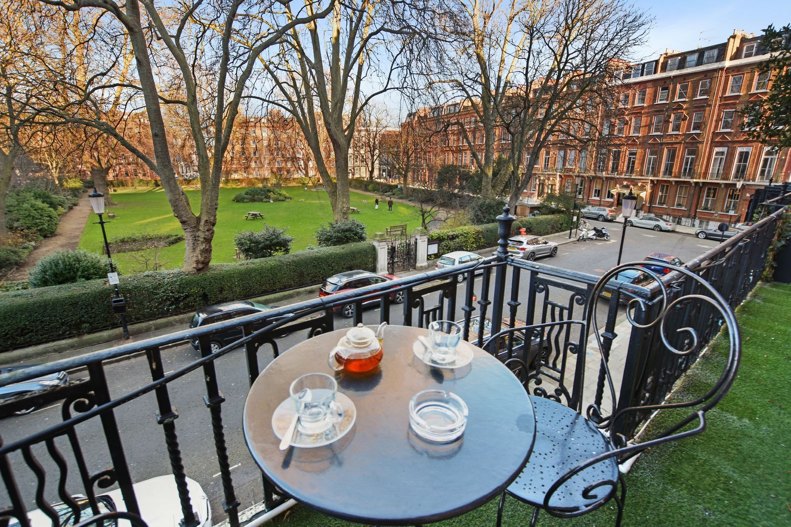 Balcony overlooking a park and street. A table with tea set, black railing, and buildings in background.