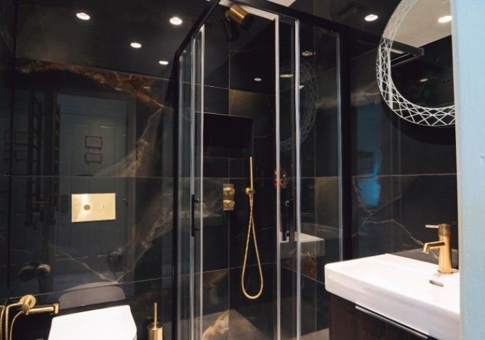Black and gold bathroom with a glass shower, toilet, and sink. Dark tile and gold fixtures.