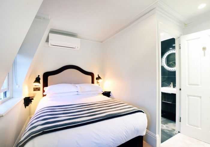 Small bedroom with bed, bedside lamps, and a door to a bathroom. White walls, air conditioner.