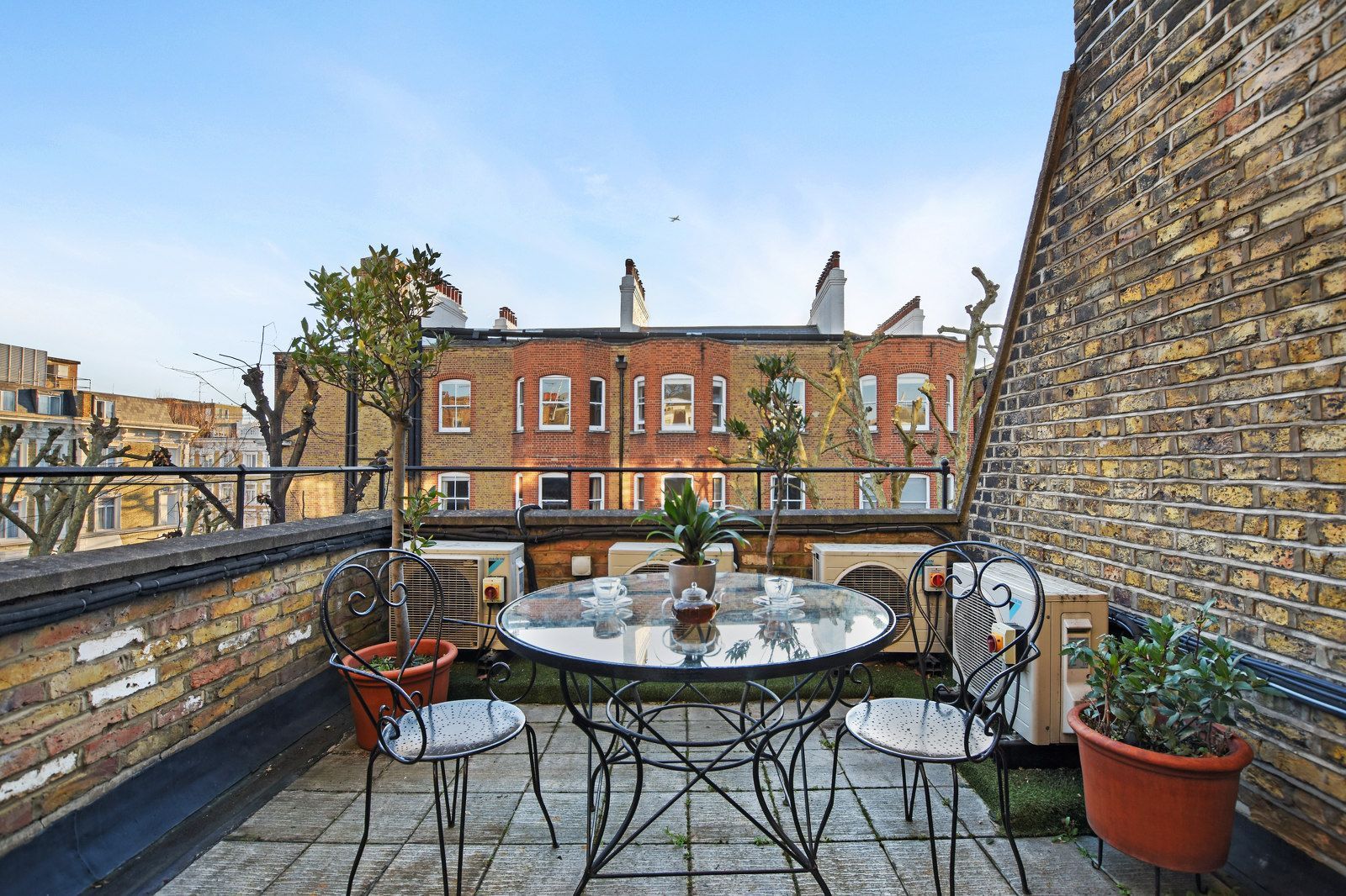 Rooftop patio with table, chairs, plants, and view of brick buildings under a blue sky.