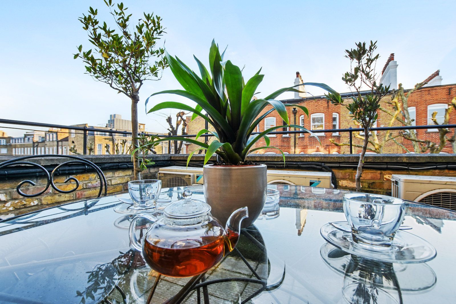 Glass table with teapot and teacups, plant in pot, cityscape background, sunny day.
