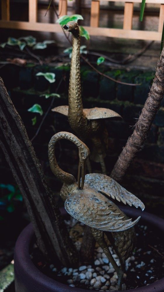 Statue of a crane with a tall, textured object in a planter with small rocks. Brick wall in background.
