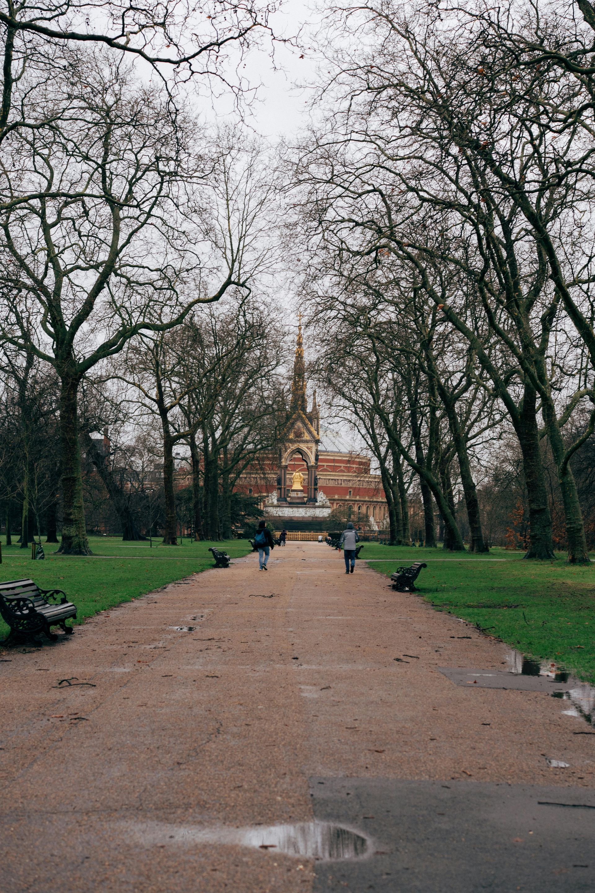 Pathway through a park, leading to a distant church. Overcast sky, bare trees, and a few people walking.