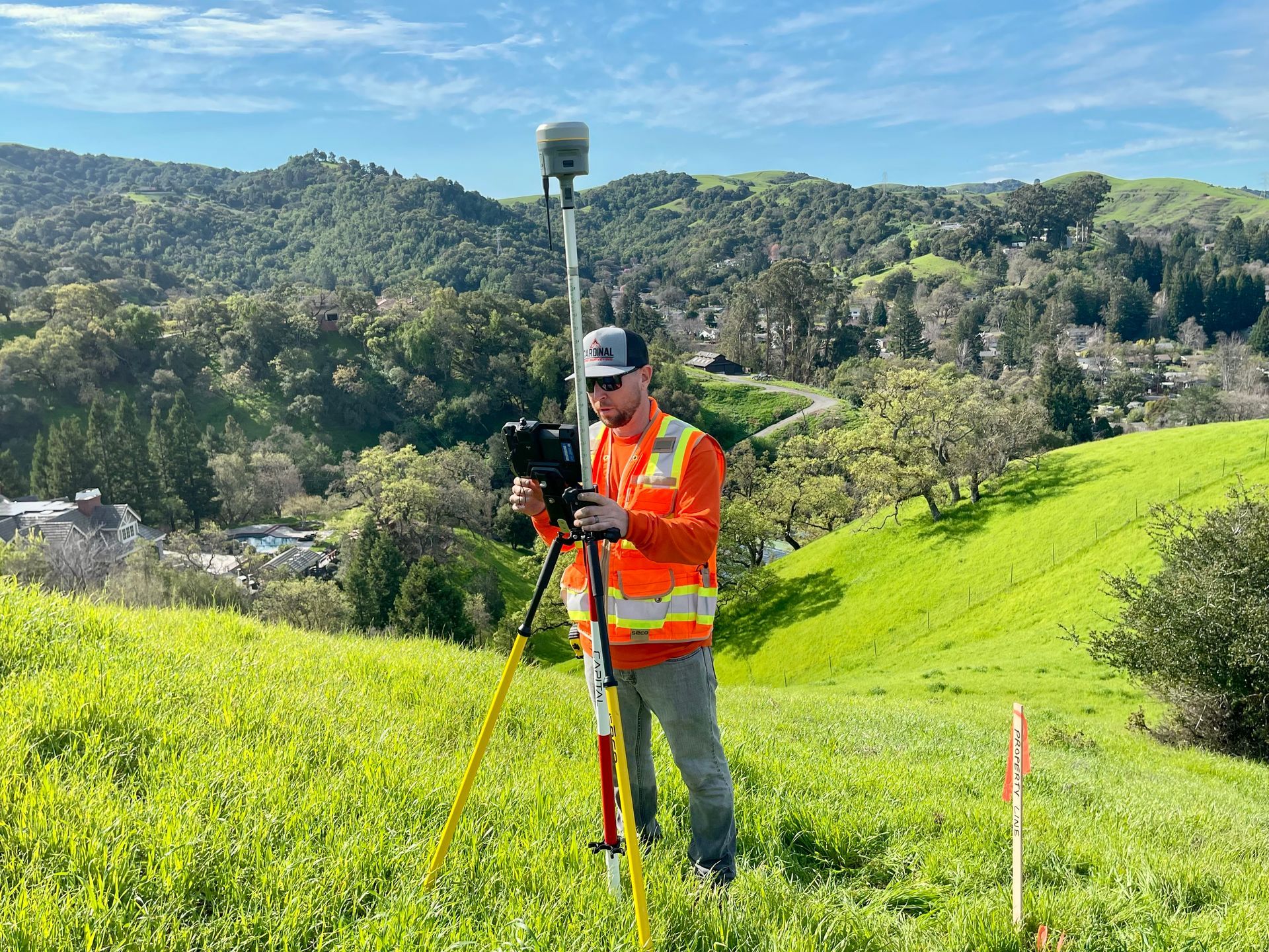Surveyor in orange vest using tripod equipment on a grassy hillside with mountains in the background
