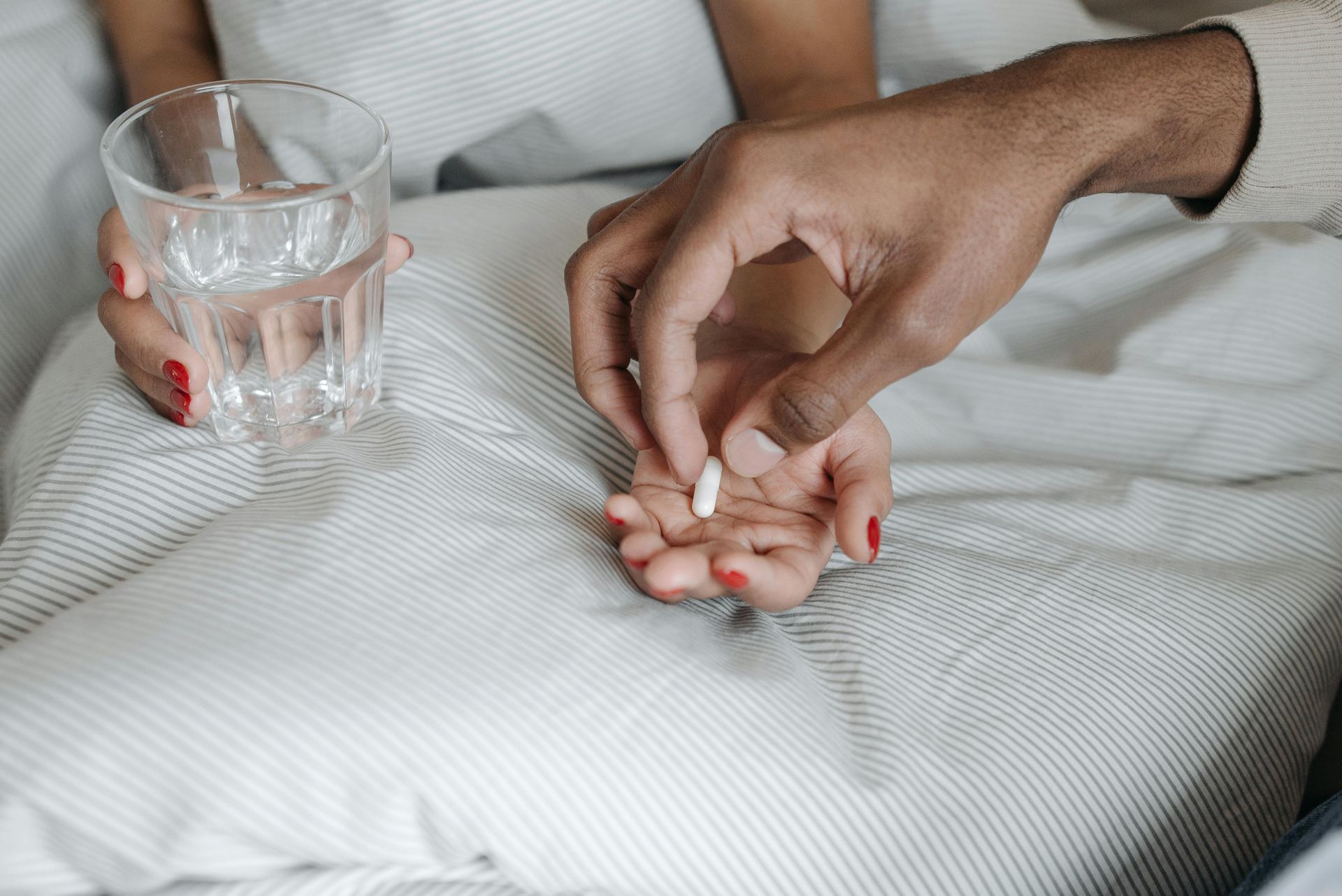 Person in bed, holding a glass of water and receiving a pill from another person.