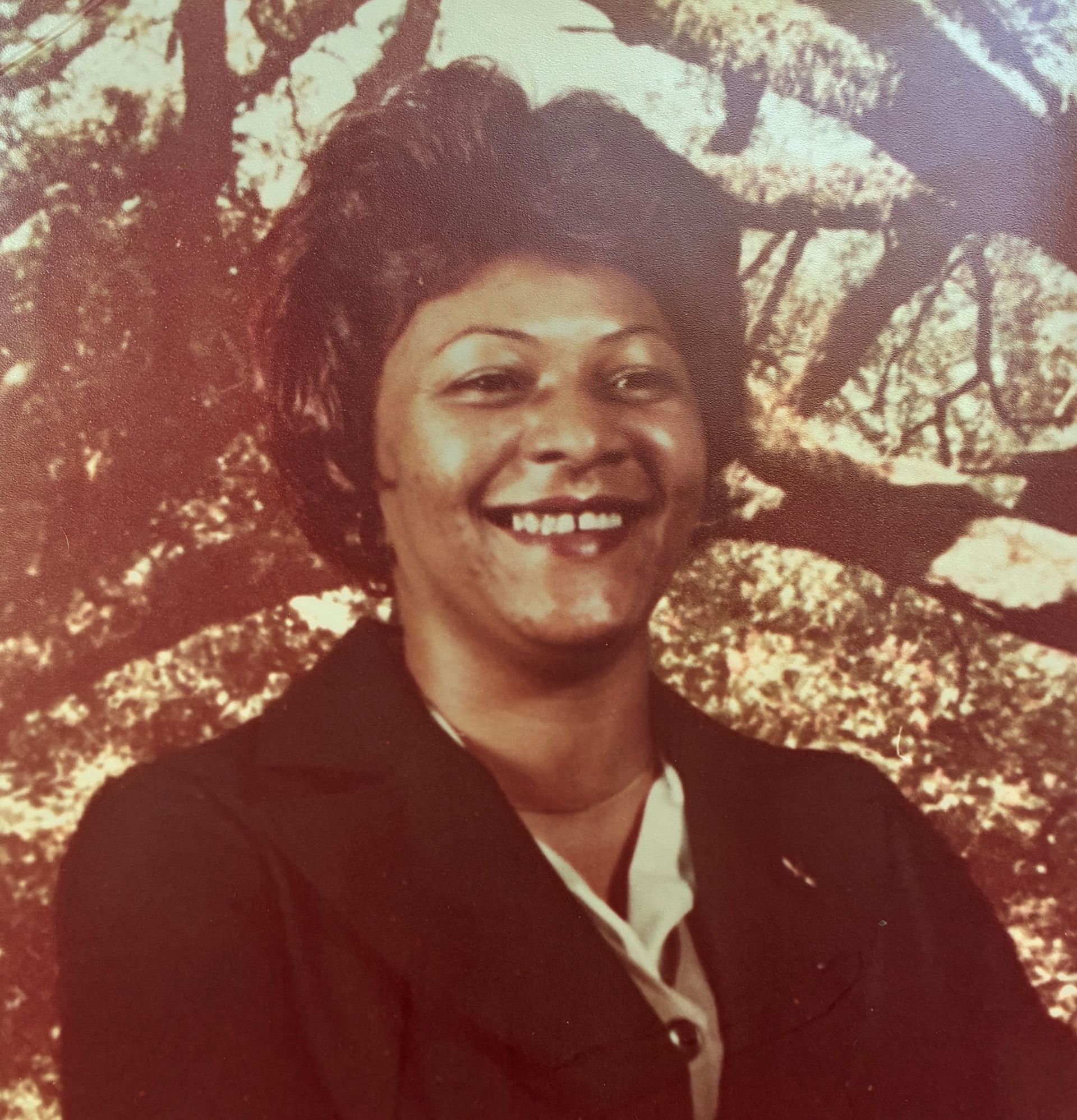 Smiling person in a dark jacket before a textured tree-patterned backdrop