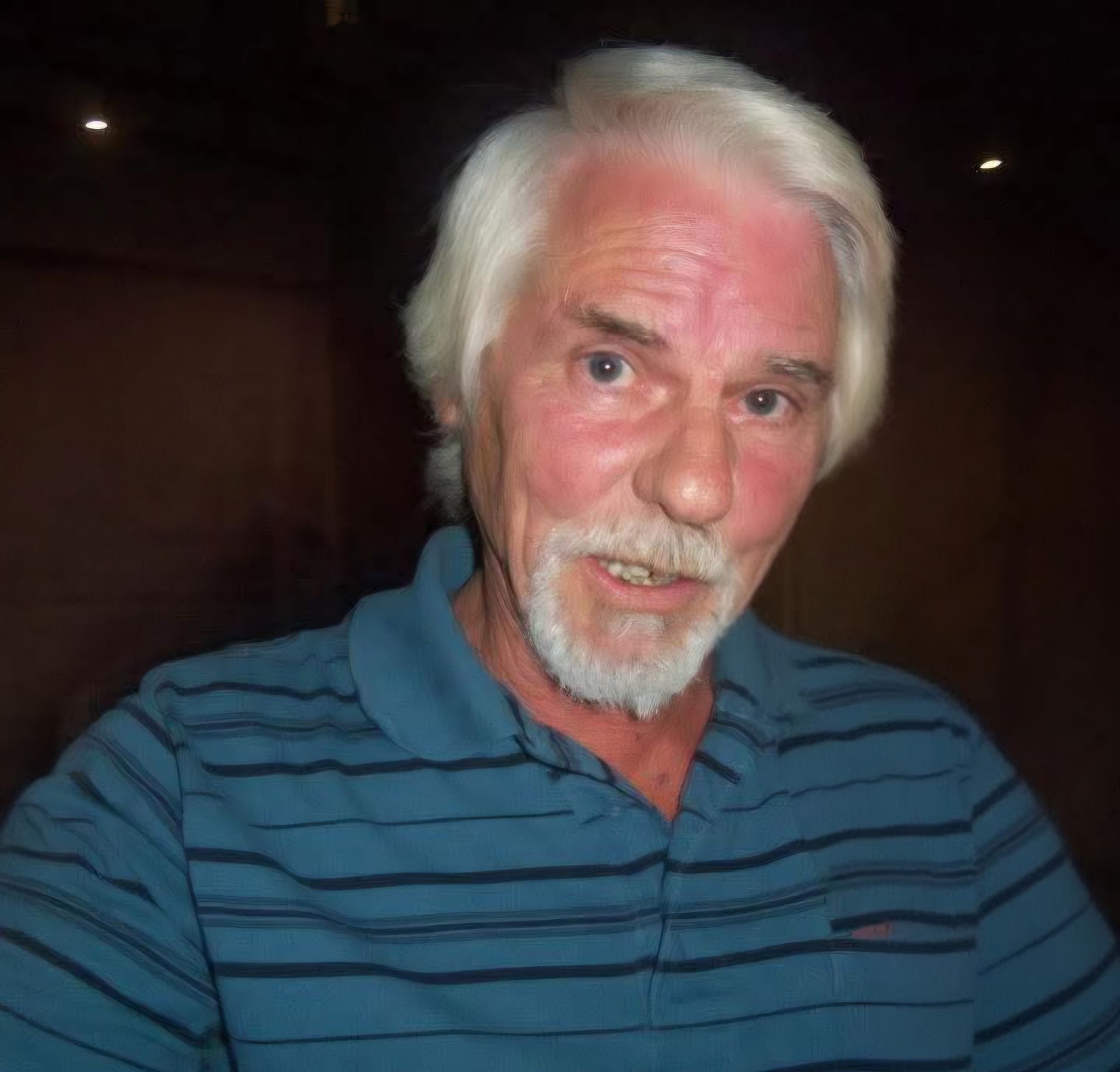 Smiling man with white hair and beard wearing a blue striped polo shirt in a dim room