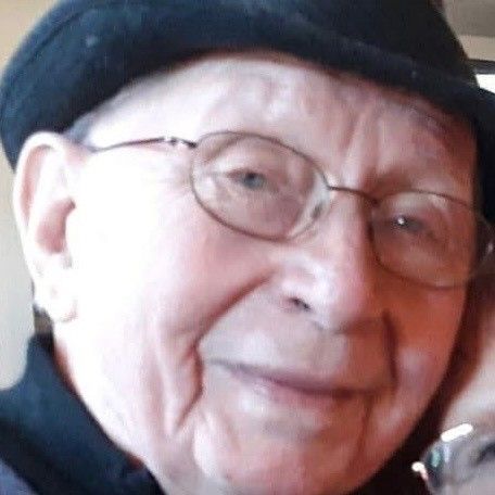 Close-up portrait of an elderly person wearing glasses and a black cap, smiling softly