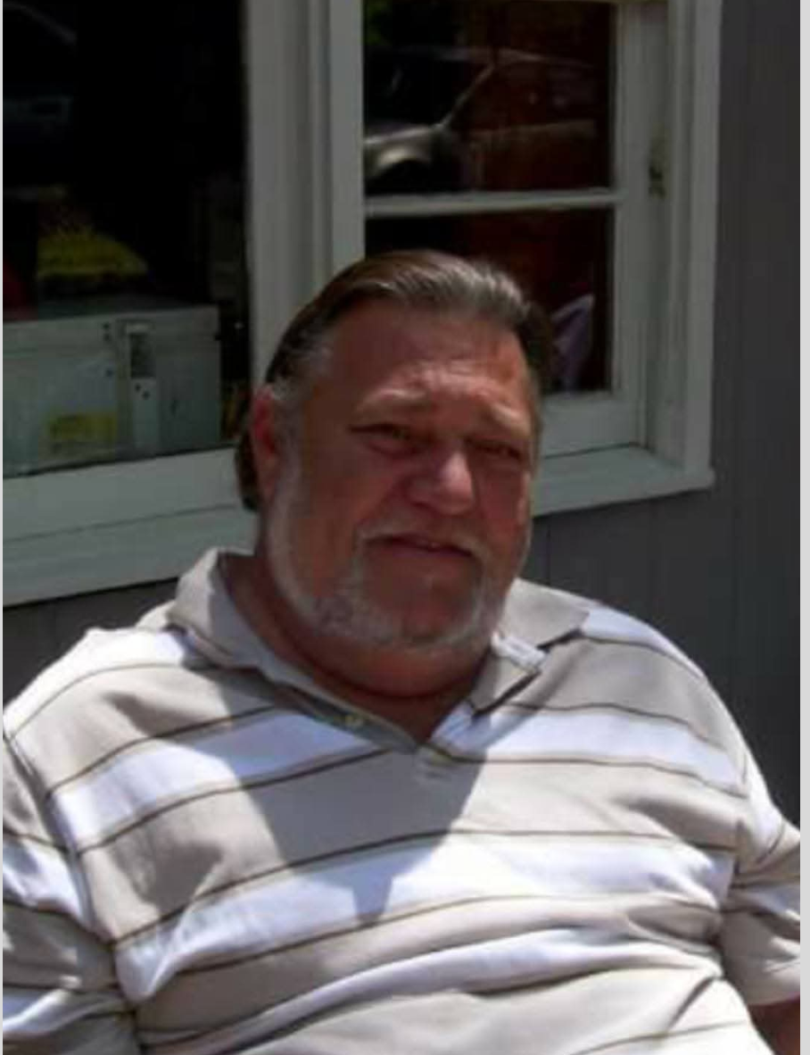Man sitting outdoors in a striped polo shirt, with a window and house siding behind him.
