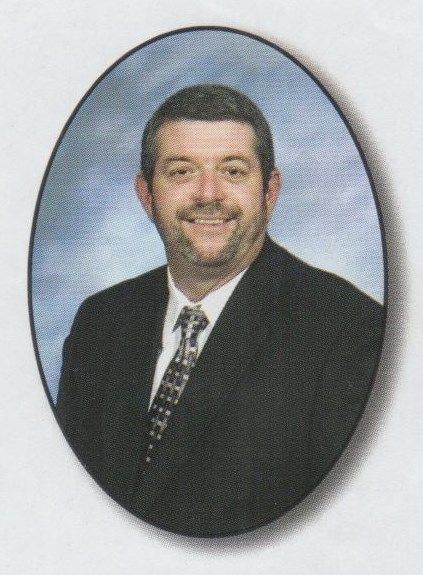 Portrait of a man in a dark suit and patterned tie, smiling against a blue oval studio backdrop