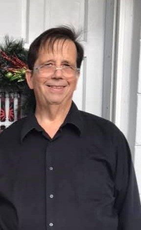 Smiling person in a black shirt standing by a doorway with a holiday wreath in the background