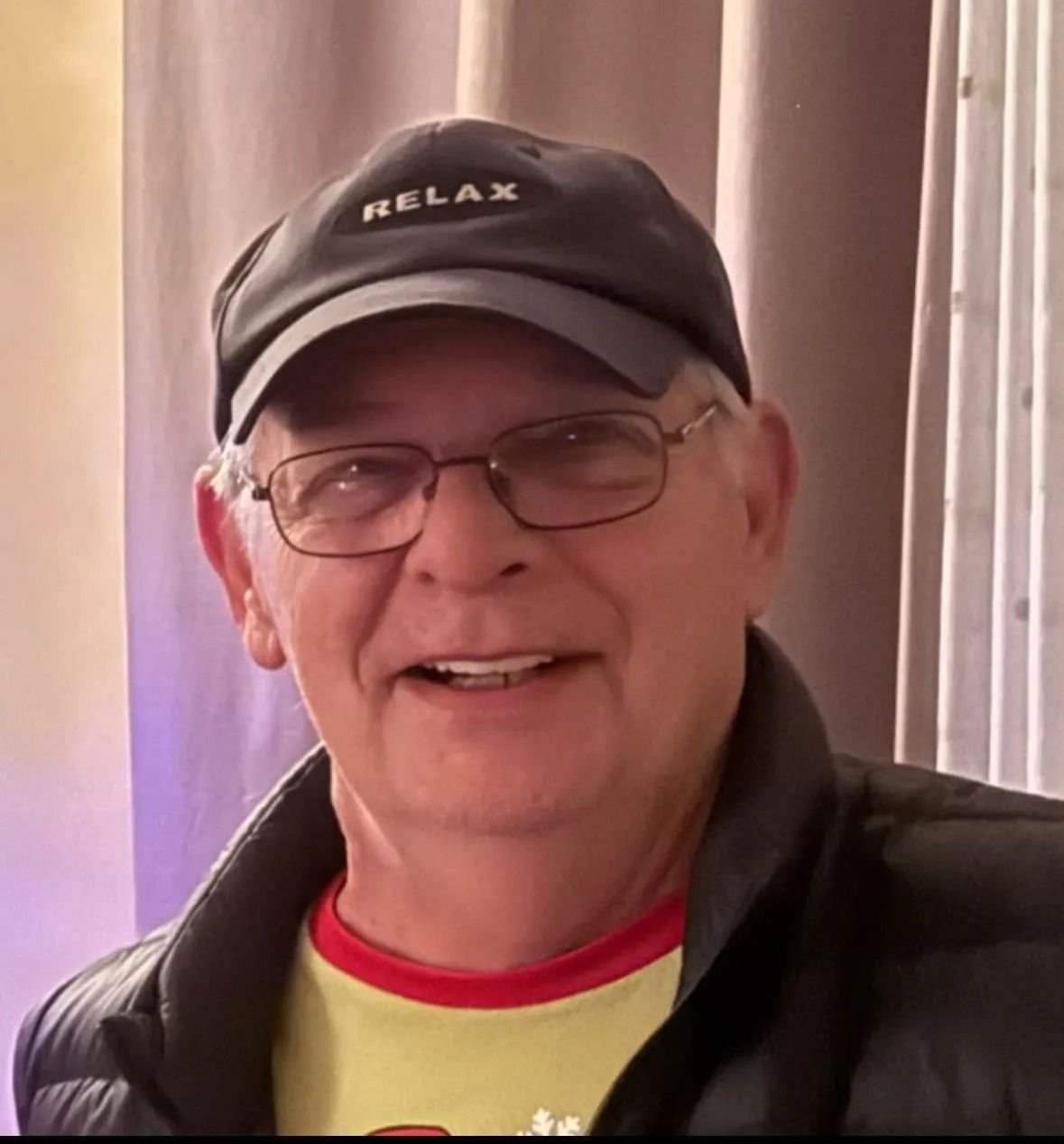 Smiling man in glasses and a black cap, wearing a jacket indoors.