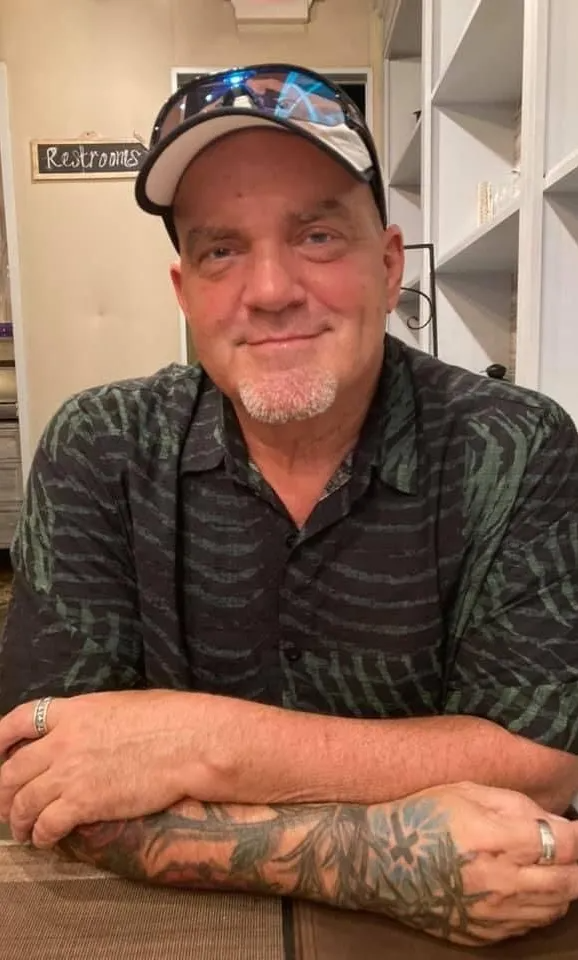 Man in a baseball cap and sunglasses leans on a counter, smiling in a kitchen.