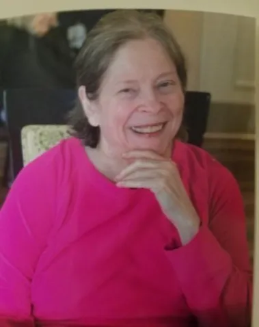 Smiling woman in a bright pink top, resting her chin on her hand indoors.