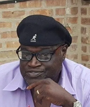 Man in a black beret and glasses, wearing a lilac shirt, resting his chin on his hand against a brick wall.