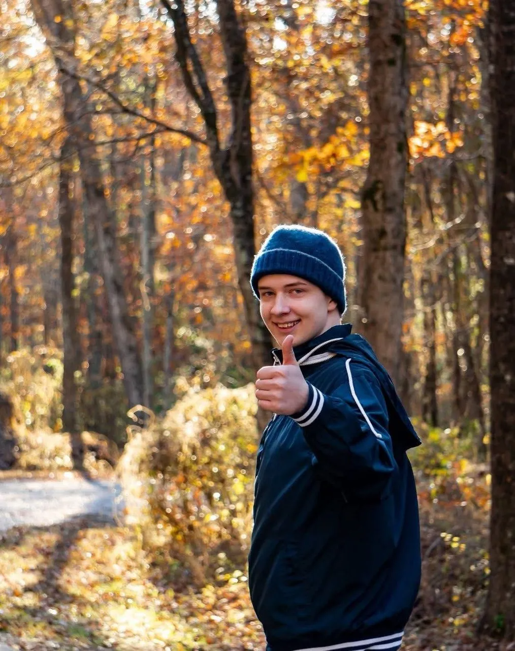 Person in a blue jacket and beanie giving a thumbs-up on a wooded trail in autumn.