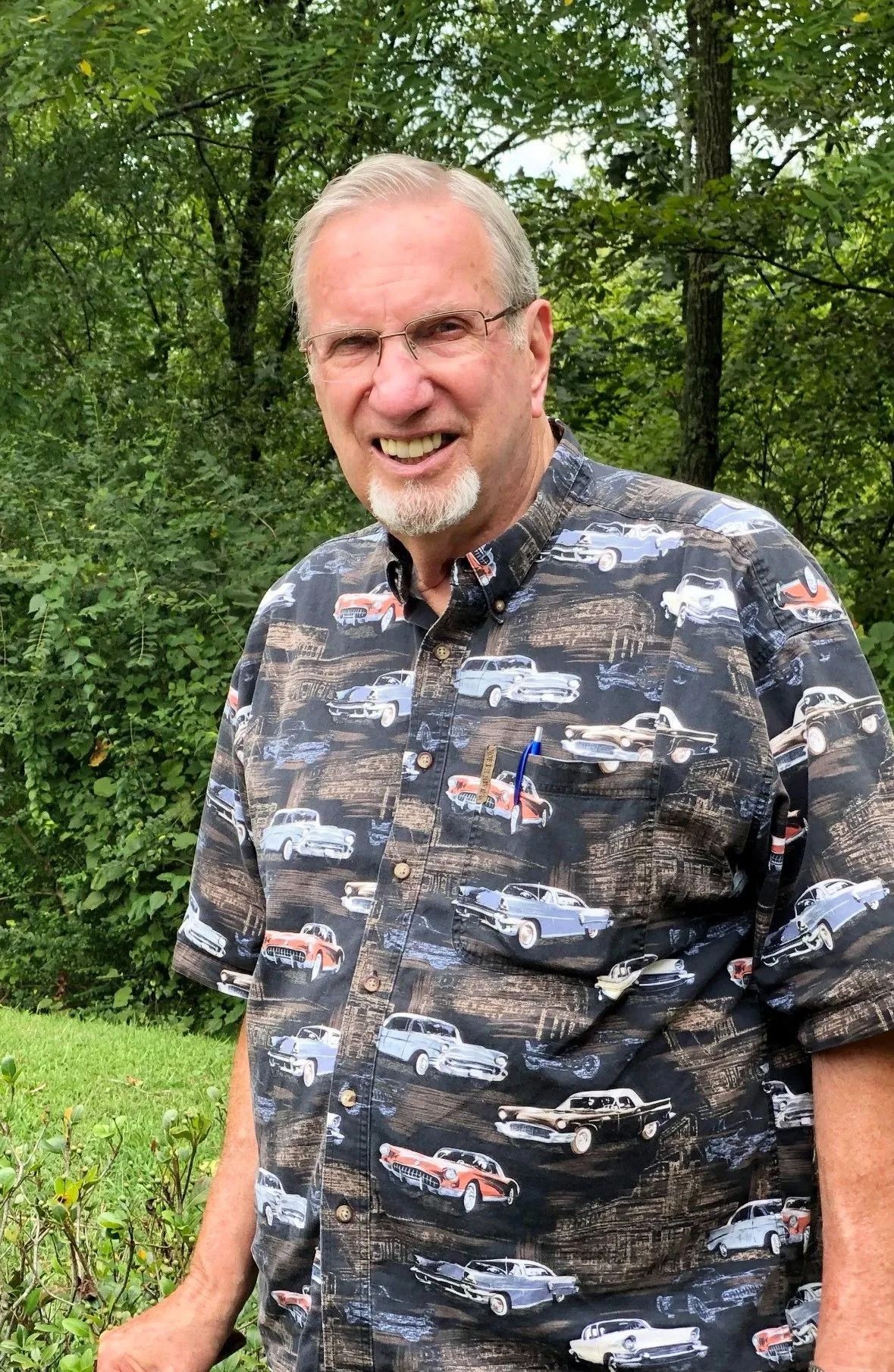Smiling man in a patterned shirt standing outdoors in a green wooded area