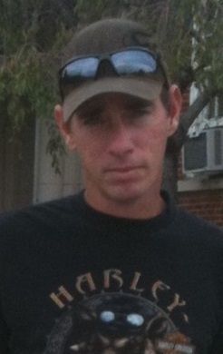 Man wearing a cap and sunglasses, standing outdoors in front of a house and trees.
