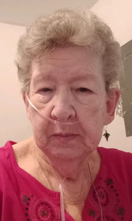 Person wearing a nasal oxygen tube and red top, looking at the camera indoors.