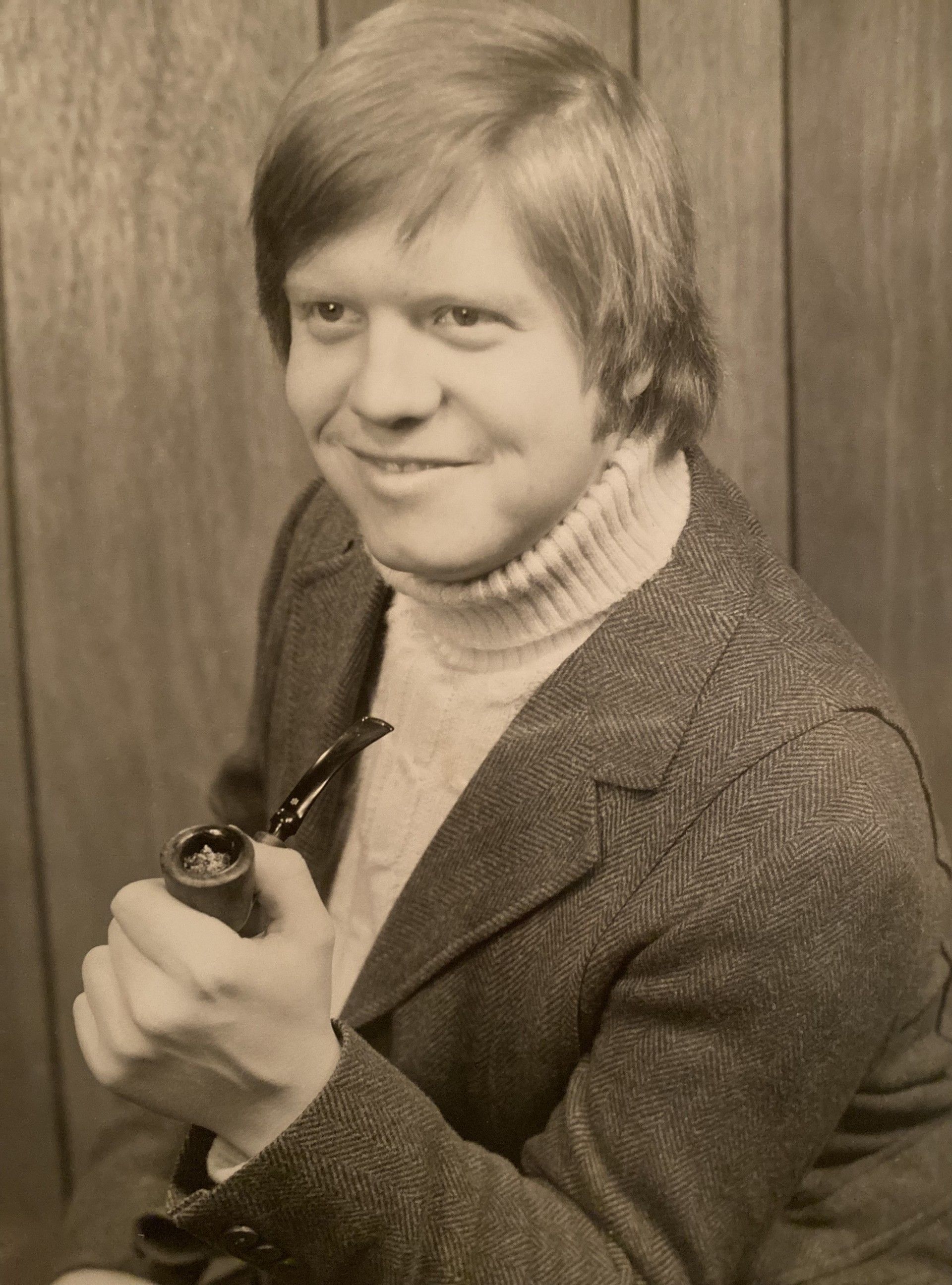 Person in a gray blazer and turtleneck holds a pipe, smiling against a wood-paneled background.