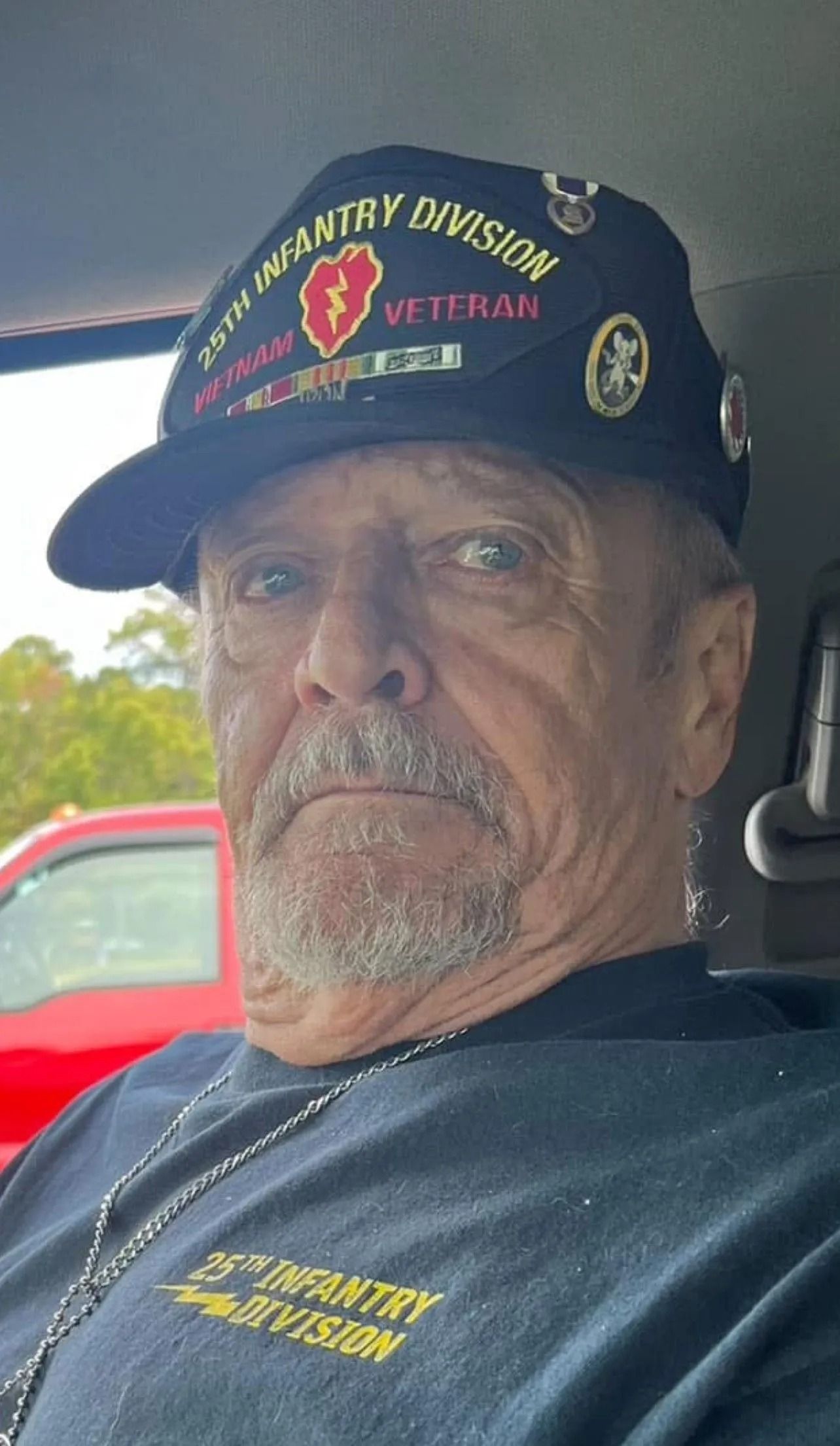 Man wearing a black cap and shirt with military patches, seated inside a vehicle with a neutral expression