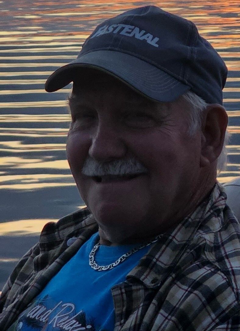Smiling man in a black cap and plaid shirt sitting by the water at sunset