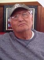 Man wearing a cap and glasses, seated in a room with wood-paneled walls and a framed picture behind him