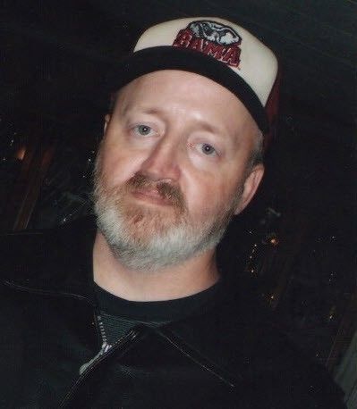 Man wearing a cream and black cap, dark jacket, and gray beard, posing indoors with a neutral expression