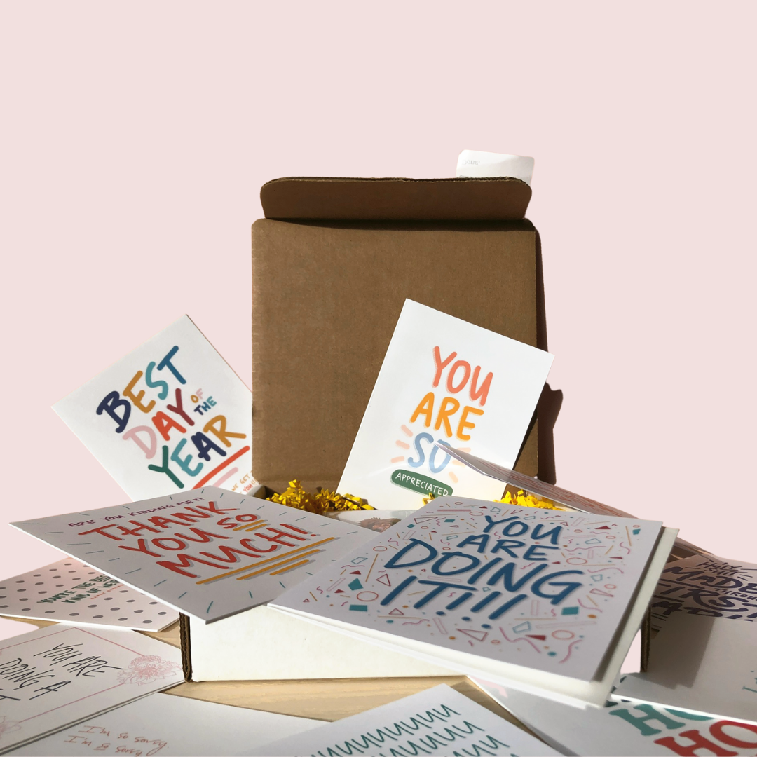 Box of colorful greeting cards scattered on a table against a pink background.